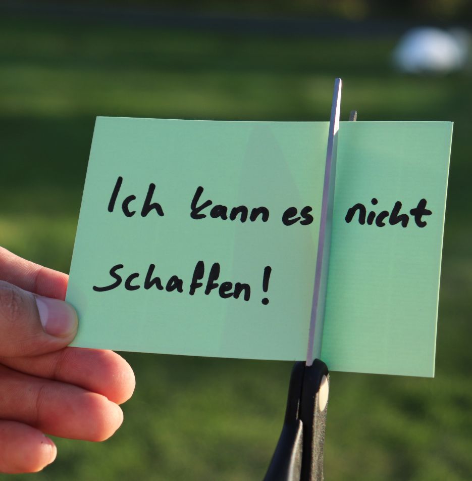 Hand hält Zettel mit Text 'Ich kann es nicht schaffen'. Eine Schere schneidet den Teil mit dem Wort 'nicht' ab.